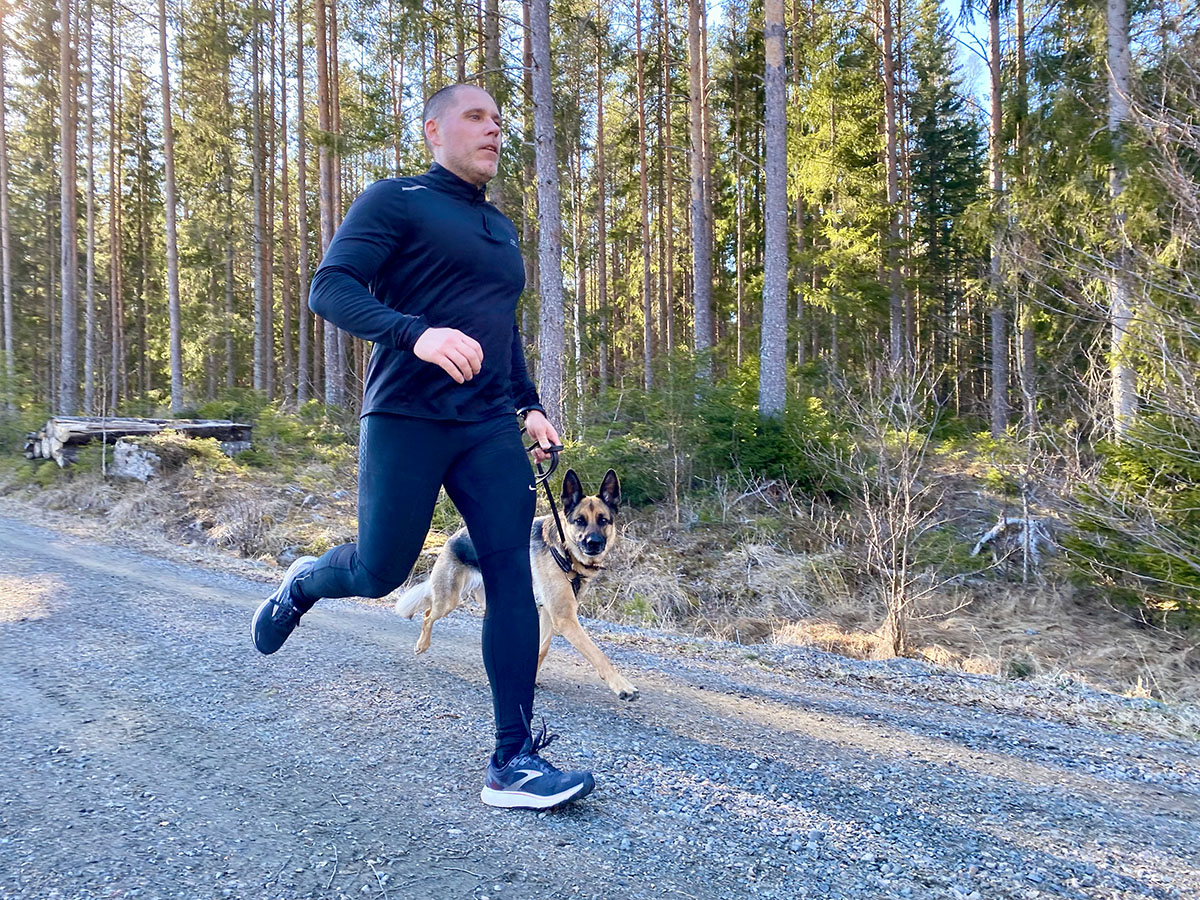 En man är ute och springer på en grusväg i skogen med sin hund